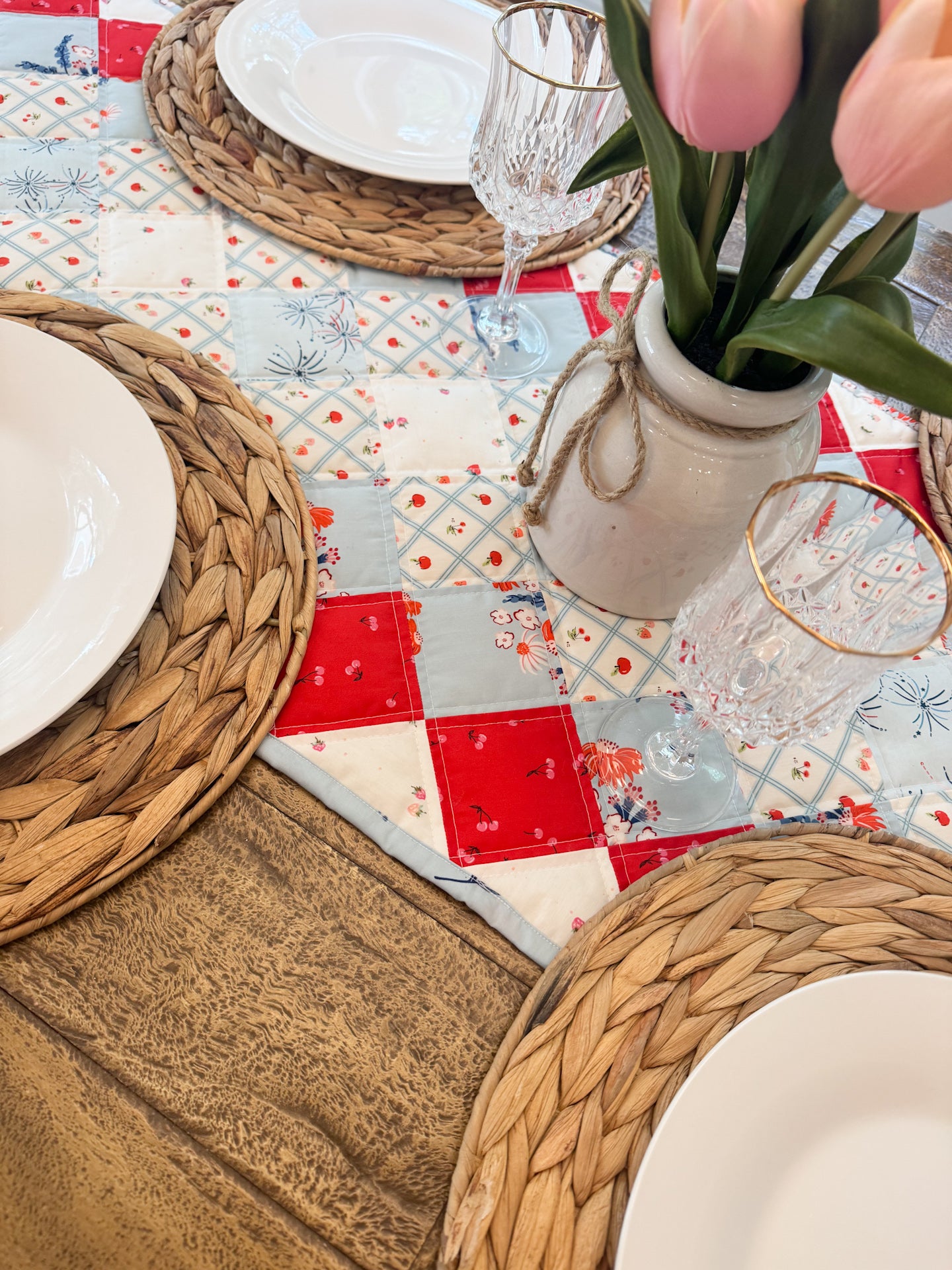 Table setting with woven placemats, white plates, and a floral centerpiece on a quilted tablecloth.
