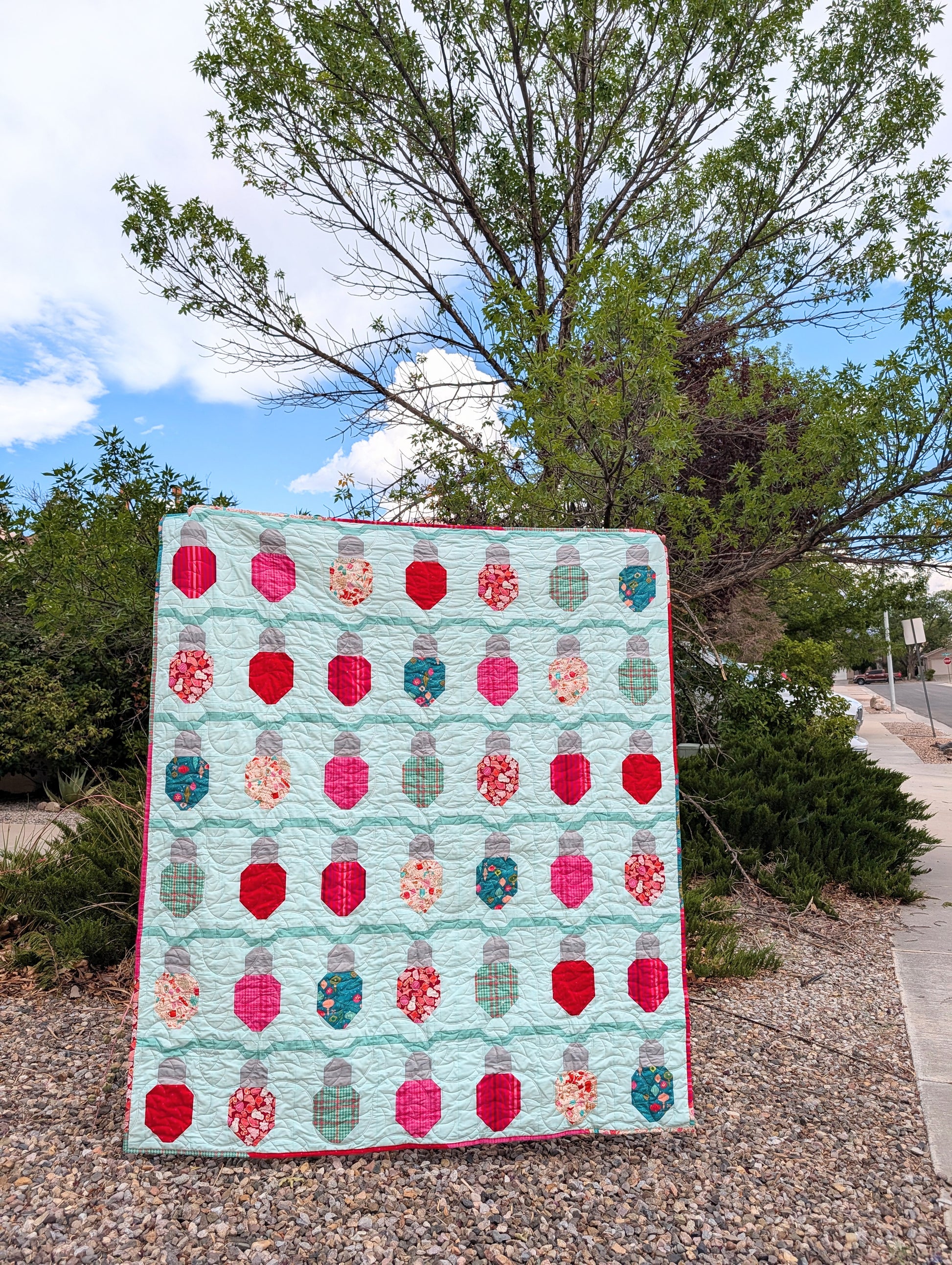 A handmade quilted patchwork blanket featuring a mint green background and rows of colorful, patterned jar-shaped patches, displayed outdoors on a gravel path with trees and sky in the background.