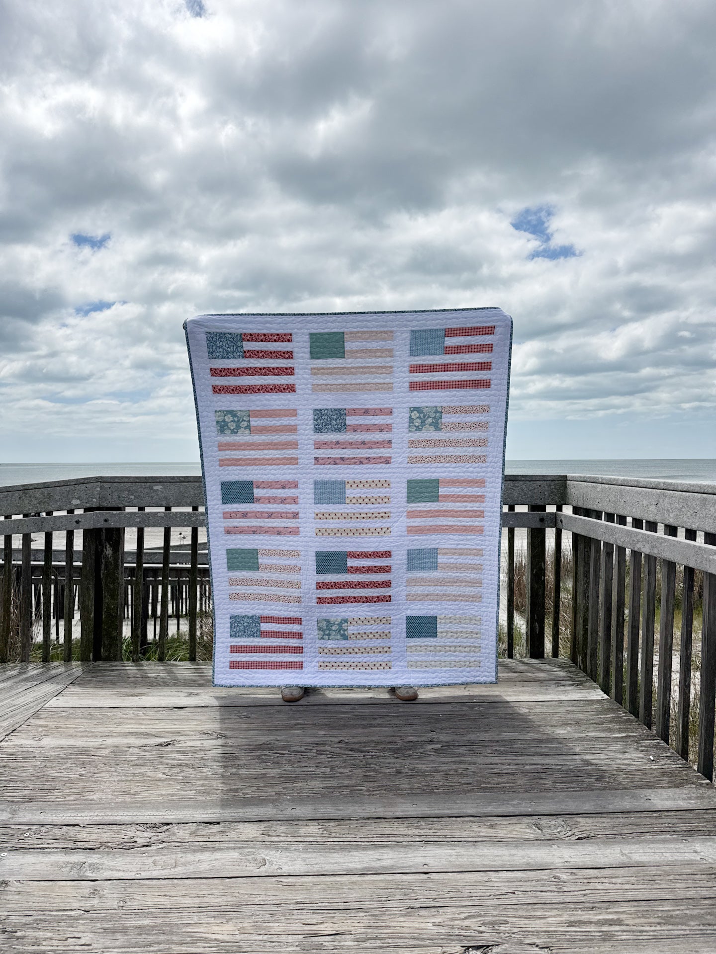Person holding a large quilt with a pattern of small flags on a wooden deck with ocean view
