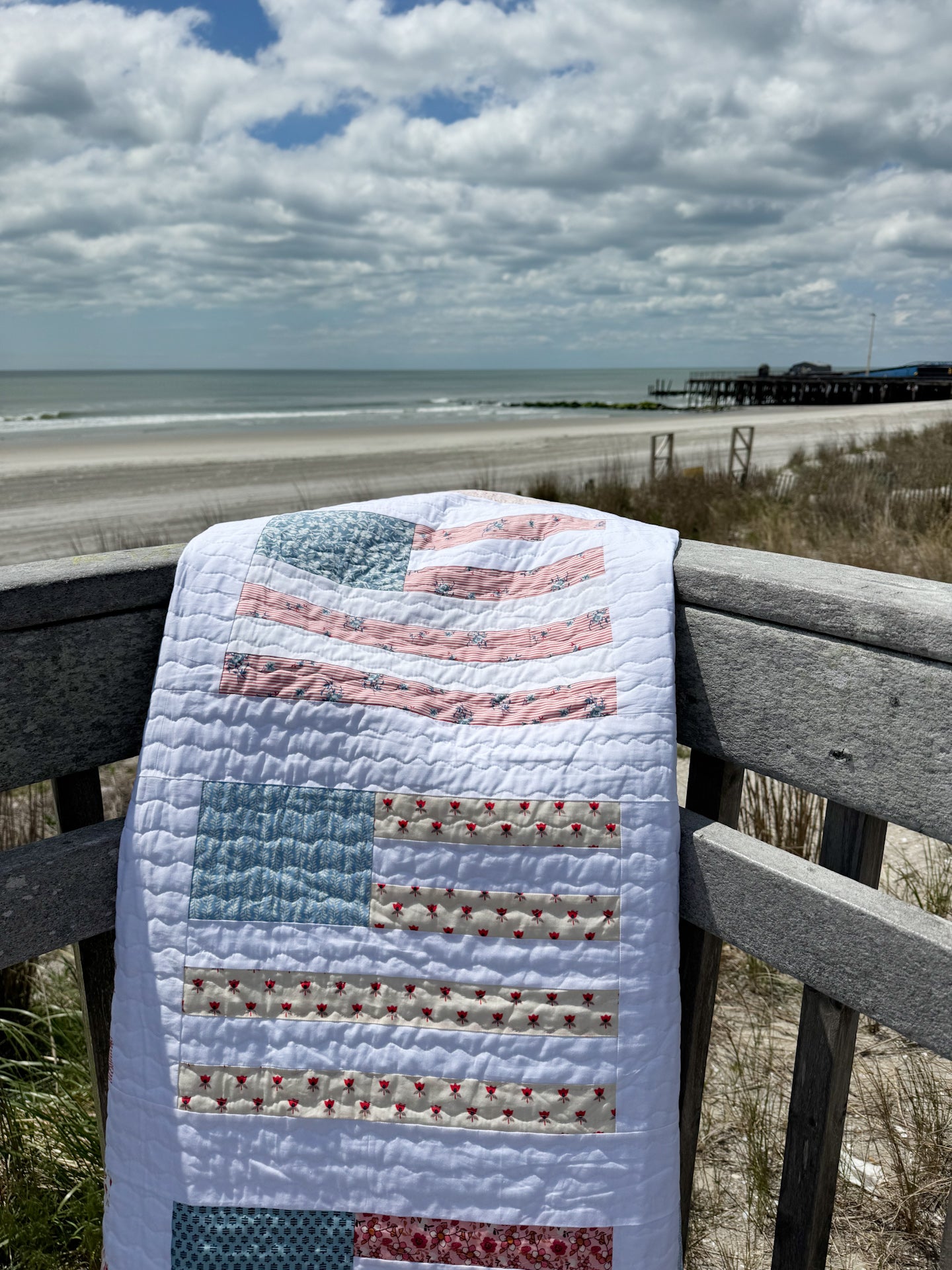 Quilt with American flag pattern draped over a railing at a beach.