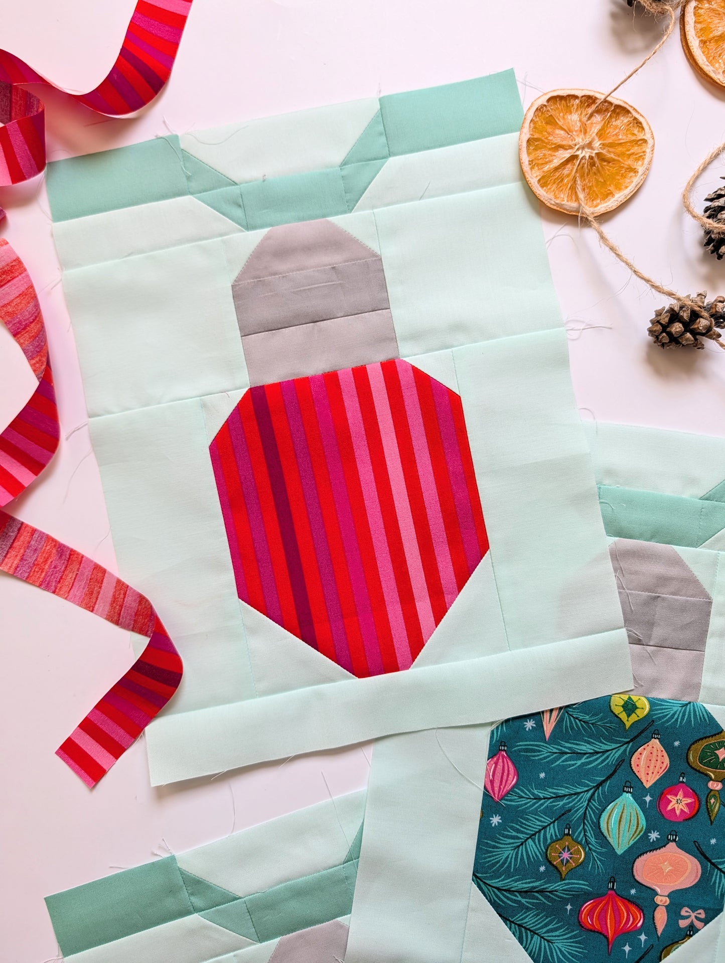 A quilted fabric block featuring a stylized Christmas ornament design with a mint green body, gray top section, and a red and pink striped ornament motif, arranged on a light pink background with ribbon, dried orange slices, and pinecones.
