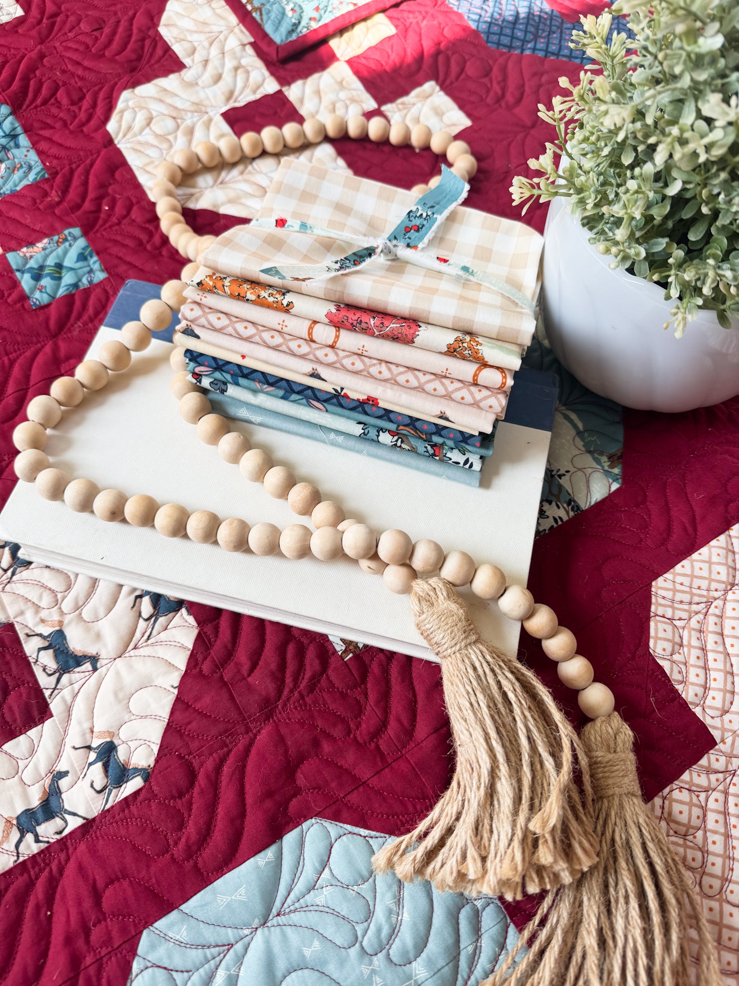 A stack of folded quilting fabrics in various patterns and colors sits on a white book, wrapped with a blue ribbon. A long wooden bead garland with tassels is draped over the stack.