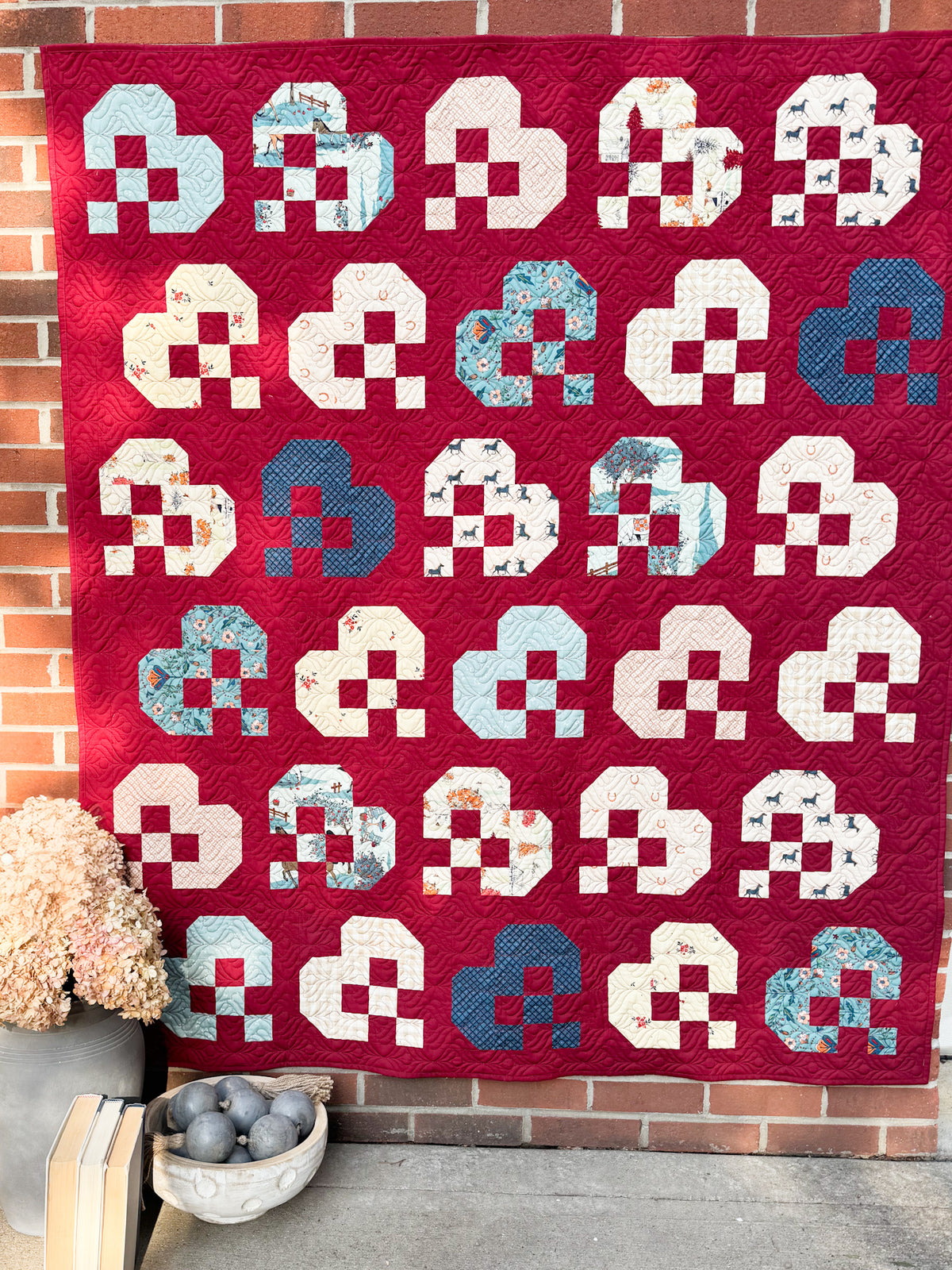 A red quilted patchwork quilt with repeating cloud-like blocks in cream, light blue, and navy, hanging against a brick wall with decorative dried flowers and a bowl of stones below.