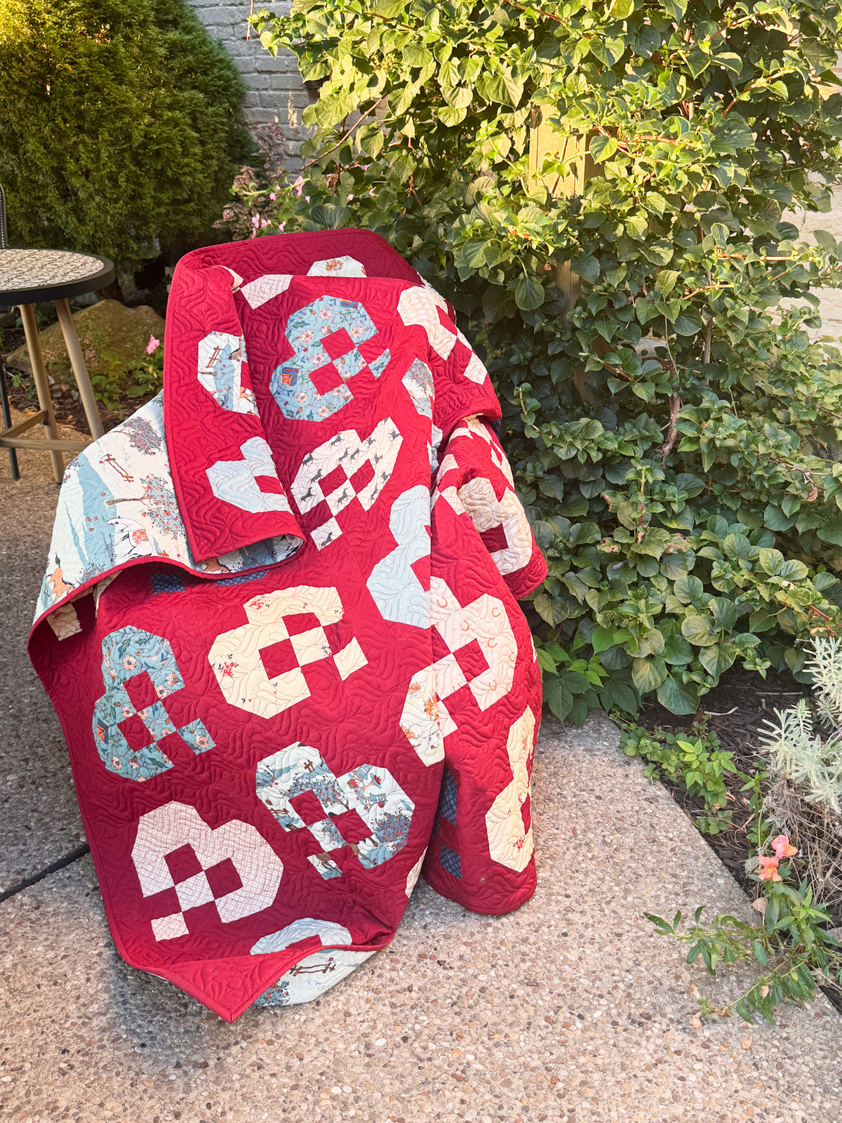 A red quilted blanket or throw with large white and light blue geometric patchwork shapes draped over a chair in an outdoor garden setting.