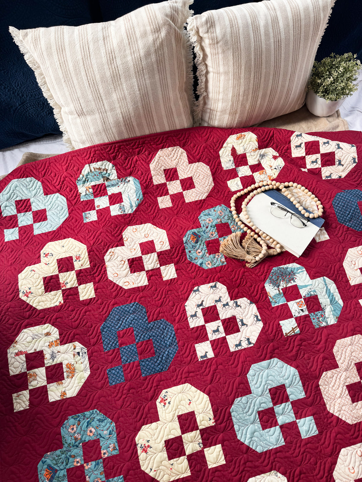 A red quilted blanket or throw with a repeating patchwork motif in cream, light blue, and navy, styled on a bed with two cream pillows and a small plant in the background.