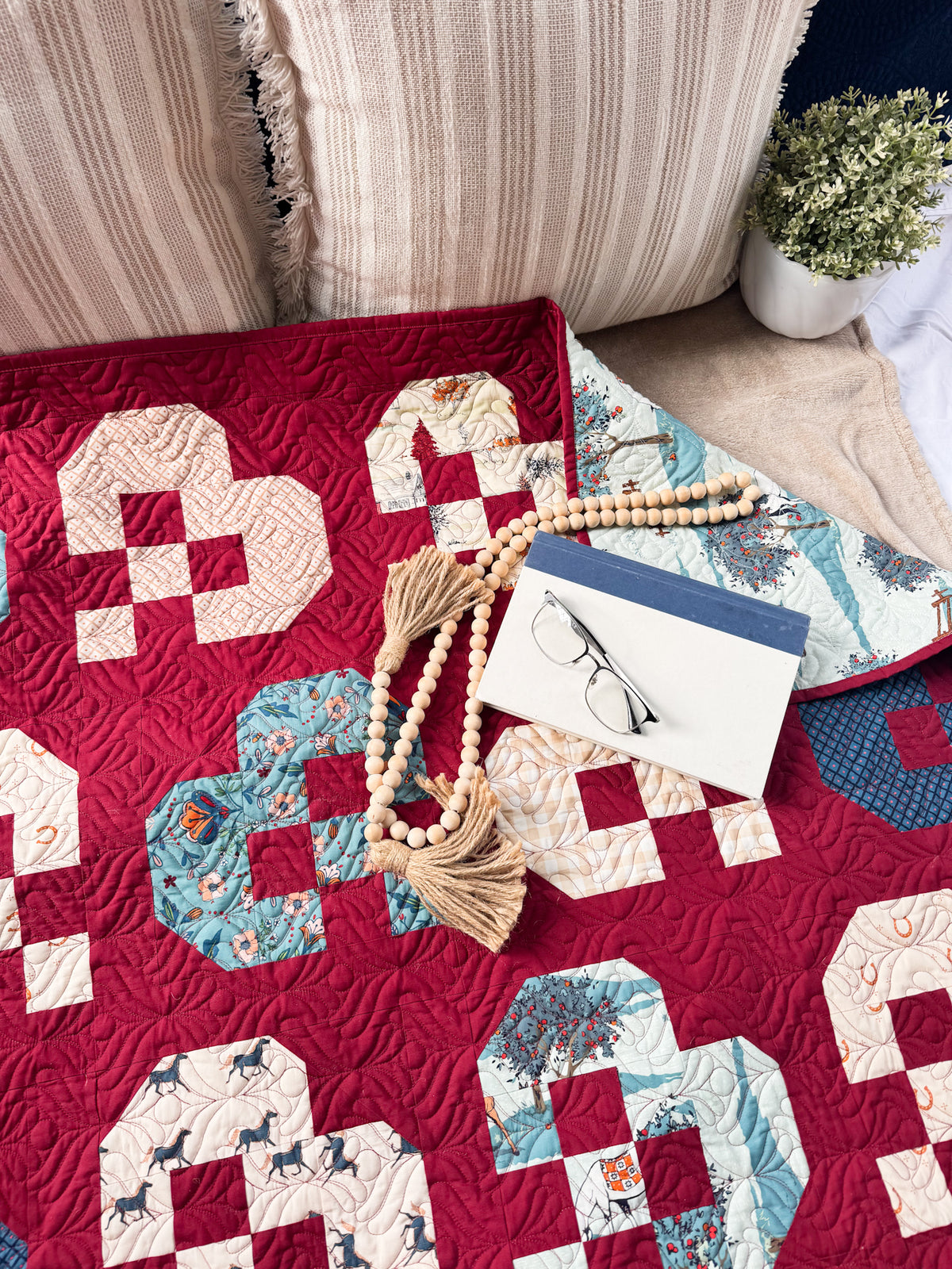 A close-up of a quilted textile with a bold red base and large cream, blue, and white patterned blocks, styled on a sofa with pillows, a wooden bead tassel necklace, a card, and a small plant.