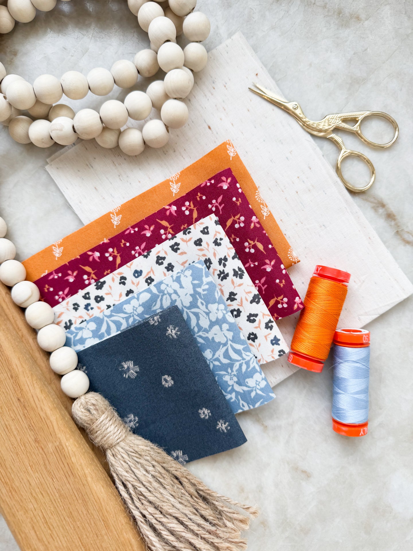 Flat lay of modern floral fabric swatches in navy, rust, and sage with orange thread and gold embroidery scissors on a marble surface.
